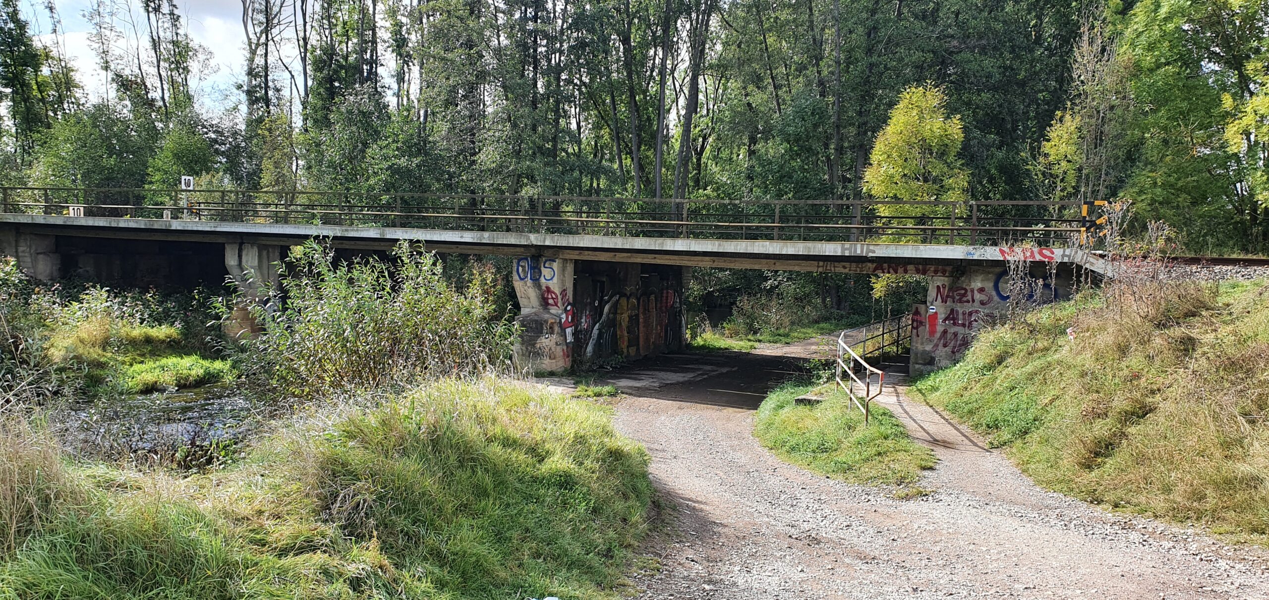 Gesamtaufnahme einer alten Bahnbrücke. Die Brücke führt über einen Fluss und einen unbefestigten Weg.