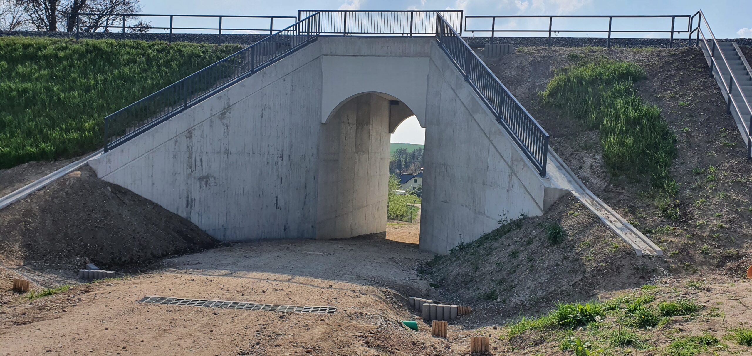 Die neue Brücke durch den hohen Bahndamm ist abgeschlossen. Die Durchfahrtshöhe hat sich deutlich vergrößert. und Bahndamm sind mit Geländern gesichert. Eine Treppe führt auf den Bahndamm hinauf.
