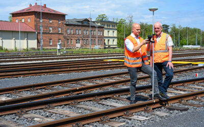Zwei Mitarbeiter der Ingenieurdienste Fenchel in Warnwesten stehen mit einem Vermessungsgerät an einer Bahnstrecke. Im Hintergrund sieht man mehrere Gleise und Häuser.