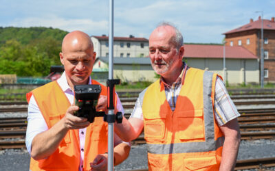 Zwei Mitarbeiter der Ingenieurdienste Fenchel in Warnwesten stehen mit einem Vermessungsgerät an einer Bahnstrecke. Im Hintergrund sieht man mehrere Gleise und Häuser.