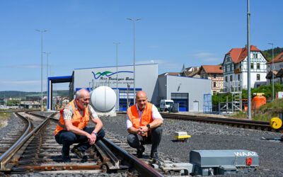 Zwei Mitarbeiter in Warnwesten hocken an einer Weiche. Im Hintergrunds ist eine Wartungshalle der Südthüringenbahn zu sehen.