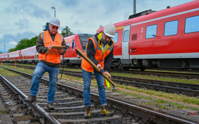 Zwei Mitarbeiter mit Warnwesten und Schutzhelmen stehen auf den Gleisen. Sie haben Werkzeuge in den Händen. Im Hintergrund stehen mehrere rote Triebwagen der Deutschen Bahn.