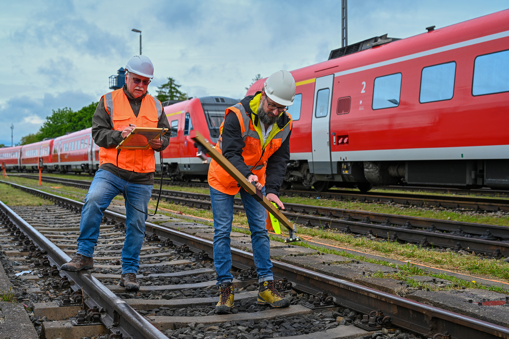 Zwei Mitarbeiter mit Warnwesten und Schutzhelmen stehen auf den Gleisen. Sie haben Werkzeuge in den Händen. Im Hintergrund stehen mehrere rote Triebwagen der Deutschen Bahn.