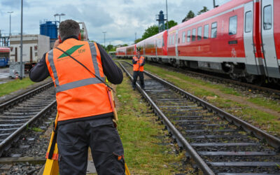 Zwei Mitarbeiter der Ingenieurdienste Fenchel stehen auf den Gleisen. Sie führen Vermessungsarbeiten durch. Auf der rechten Seite sind mehrere rote Triebwagen zu sehen.