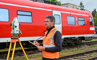 Ein Mitarbeiter mit Warnweste steht auf den Gleisen. Er hat ein Messgerät vor sich. Im Hintergrund steht ein roter Triebwagen der Deutschen Bahn.