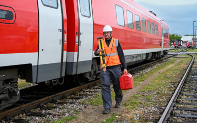 Ein Mitarbeiter mit Vermessungsausrüstung, Warnweste und Helm läuft an einem Gleis entlang. Auf dem Gleis steht ein roter Triebwagen der Deutschen Bahn. Im Hintergrund sind weitere rote Triebwagen zu sehen.