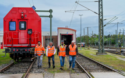 Vier Mitarbeiter laufen auf die Kamera zu. Sie tragen Warnwesten. Sie kommen aus Richtung eines Bahn-Werkes. Rechts und Links sind Gleise zu sehen. Ein Bauzug steht auf dem linken Gleis.