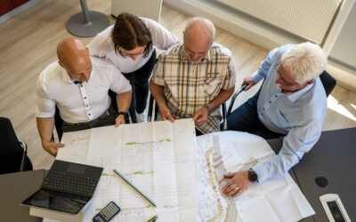 Vier Mitarbeiter sitzen und stehen an einem Tisch. Auf dem Tisch liegen mehrere Pläne, ein Laptop und ein Taschenrechner.