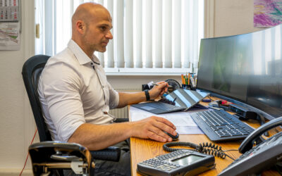 Ein Mitarbeiter sitzt an einem Schreibtisch. Auf dem Schreibtisch ist ein großer Bildschirm, ein Laptop und ein Telefon.