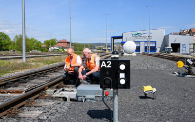Zwei Mitarbeiter in Warnwesten hocken an einer Weiche. Im Hintergrunds ist eine Wartungshalle der Südthüringenbahn zu sehen.