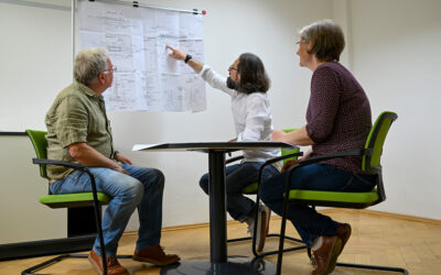Drei Menschen sitzen an einem kleinen Tisch und schauen auf einen Bauplan, der im Hintergrund hängt.