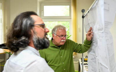 Zwei Menschen stehen vor einer Planzeichnung. Im Hintergrund ist ein Fenster.