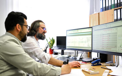 Zwei Mitarbeiter sitzen an einem Schreibtisch und schauen auf Planungen, welche auf zwei Bildschirmen zu sehen sind.